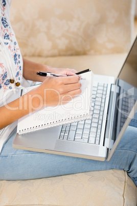 Close up of woman writing on notepad with laptop on her legs