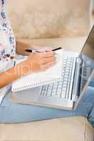 Close up of woman writing on notepad with laptop on her legs