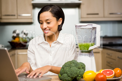 Smiling brunette using laptop and preparing smoothie