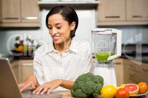 Smiling brunette using laptop and preparing smoothie