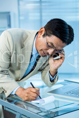 Overwhelmed asian businessman answering the phone and writing