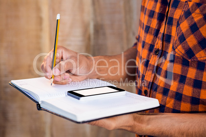 Mid section of man with smartphone writing on book in office