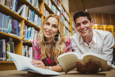 Smiling classmates reading book while leaning on bookshelves