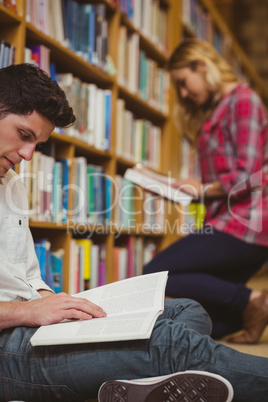 Smiling classmates reading book while leaning on bookshelves