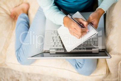 Mid section of woman writing on notepad with laptop on her legs