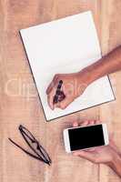 Businessman writing in notebook while holding smartphone in offi
