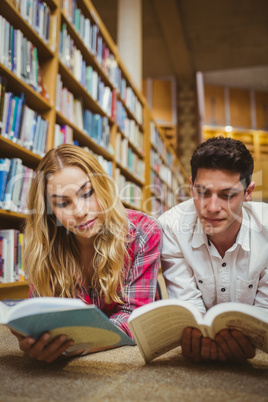 Smiling classmates reading book while leaning on bookshelves