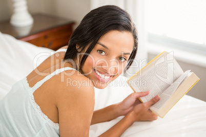 Smiling brunette holding a book on bed