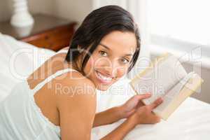 Smiling brunette holding a book on bed