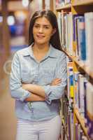 Smiling brunette student standing next to bookshelves with arms