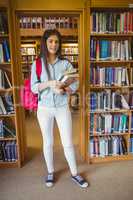 Smiling brunette student standing next to bookshelves while hold