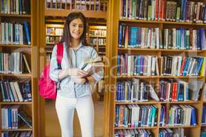 Smiling brunette student standing next to bookshelves while hold