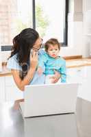 Smiling brunette holding her baby and using laptop on phone call