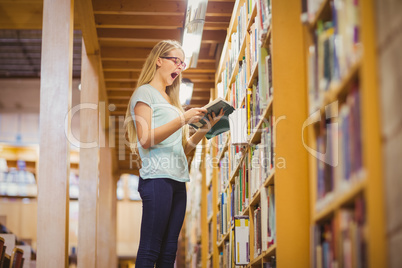 Blonde student reading book next to bookshelf