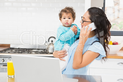 Smiling brunette holding her baby and using laptop on phone call