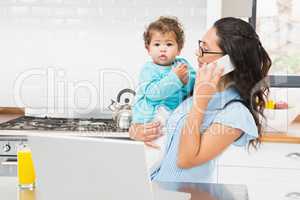 Smiling brunette holding her baby and using laptop on phone call