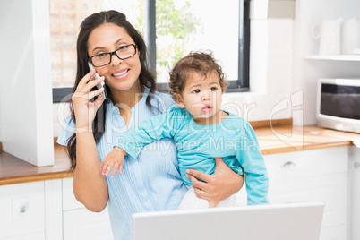 Smiling brunette holding her baby and using laptop on phone call