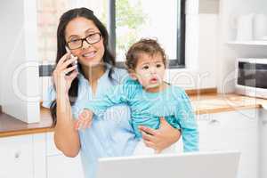 Smiling brunette holding her baby and using laptop on phone call