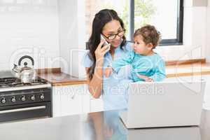 Smiling brunette holding her baby and using laptop on phone call