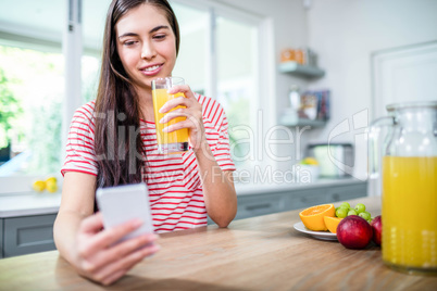 Smiling brunette using smartphone and drinking orange juice