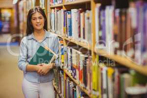 Smiling brunette student standing next to bookshelves while hold