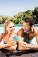 Happy couple leaning on pool edge and toasting with cocktails