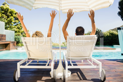 Rear view of couple raising hands and lying on deck chairs