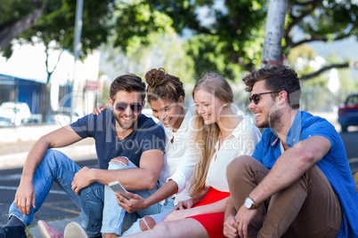 Hip friends looking at smartphone and sitting on sidewalk