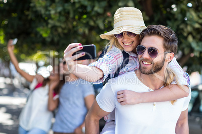 Hip man giving piggy back to his girlfriend and taking selfie