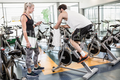 Female trainer looking at stopwatch and man using exercise bike