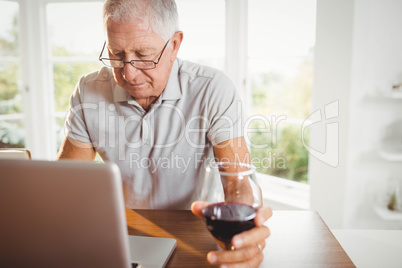 Focused senior man using laptop and drinking wine