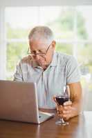 Focused senior man using laptop and drinking wine