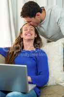 Woman using laptop and man kissing on her forehead