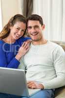 Young couple smiling face to face on sofa and using laptop
