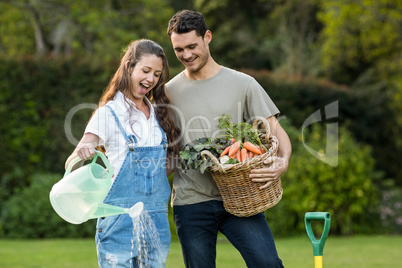 Woman watering a plants while man holding basket of vegetables