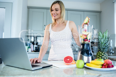 Pretty blonde woman preparing a smoothie with recipe on laptop