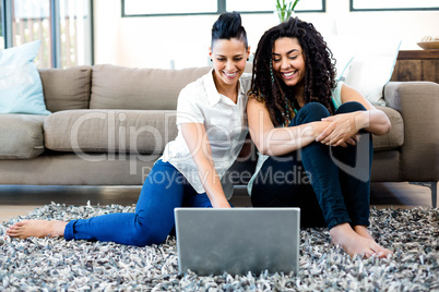 Smiling lesbian couple sitting on rug and using laptop