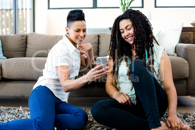 Smiling lesbian couple sitting on rug and looking at their mobil