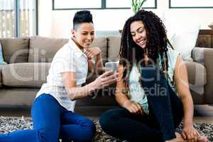 Smiling lesbian couple sitting on rug and looking at their mobil