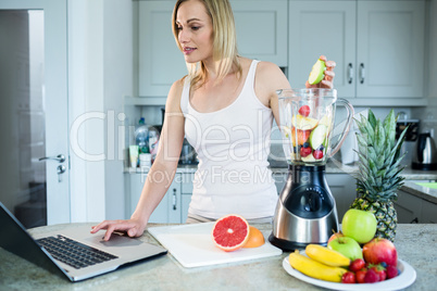 Pretty blonde woman preparing a smoothie with recipe on laptop