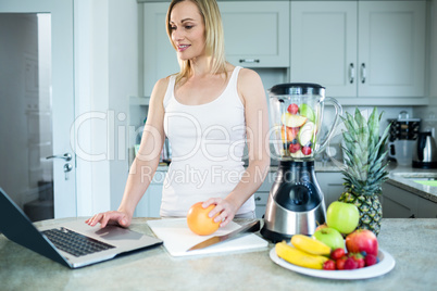 Pretty blonde woman preparing a smoothie with recipe on laptop