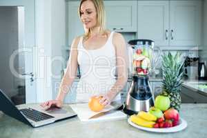 Pretty blonde woman preparing a smoothie with recipe on laptop