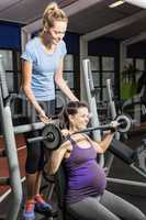Smiling trainer helping pregnant woman lifting barbell