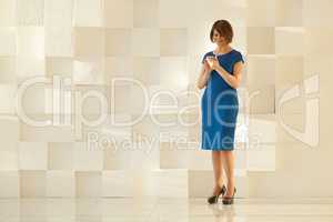 Woman in blue dress standing against modern wall while looking at smartphone