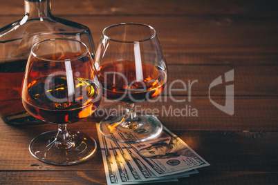 Two glasses of cognac and bottle, with wad of money on the wooden table.