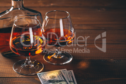 Two glasses of cognac and bottle, with wad of money on the wooden table.
