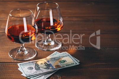 Two glasses of cognac with wad of money on the wooden table.