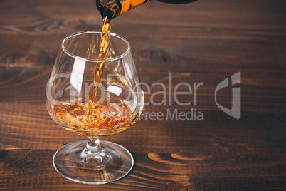 Pouring cognac or whiskey from the bottle into the glass against wooden background