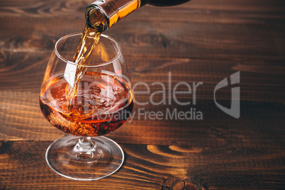 Pouring cognac or whiskey from the bottle into the glass against wooden background