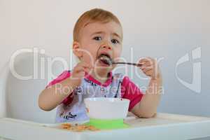 Little smiling baby sits at highchair and eats porridge on plate.